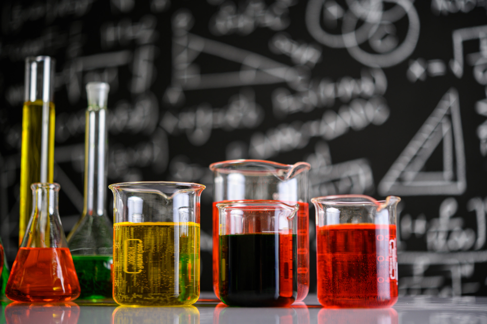 Pahare și recipiente de laborator cu soluții colorate, pe fundalul unei table cu formule chimice