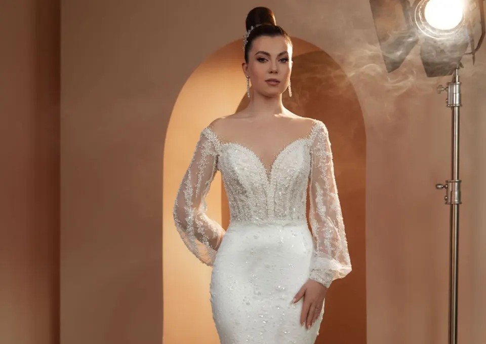 Model purtând o rochie de mireasă albă Adora Bridal, cu croială sirenă, corset cu aplicații strălucitoare, decolteu adânc și mâneci lungi transparente, brodate.